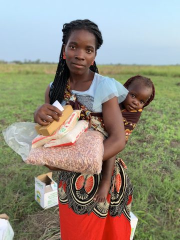 Nainen sylisään ruokapusseja ja saippuaa, kantaa liinassa lasta selässä.