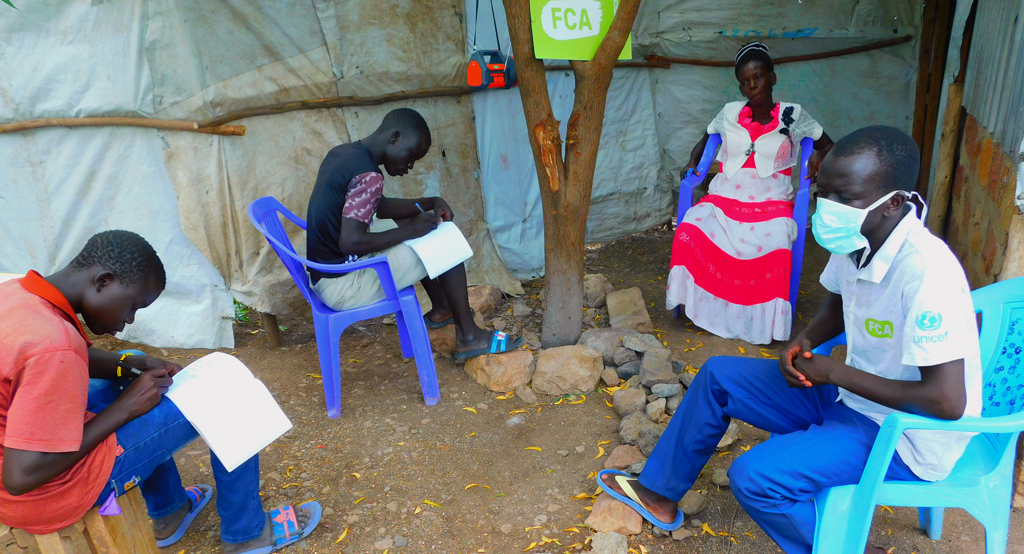 Two students and two adults sitting in plastic chairs in the yard, listening to a radio lessons from a small radio hanged on tree branch and writing in their exercise books.