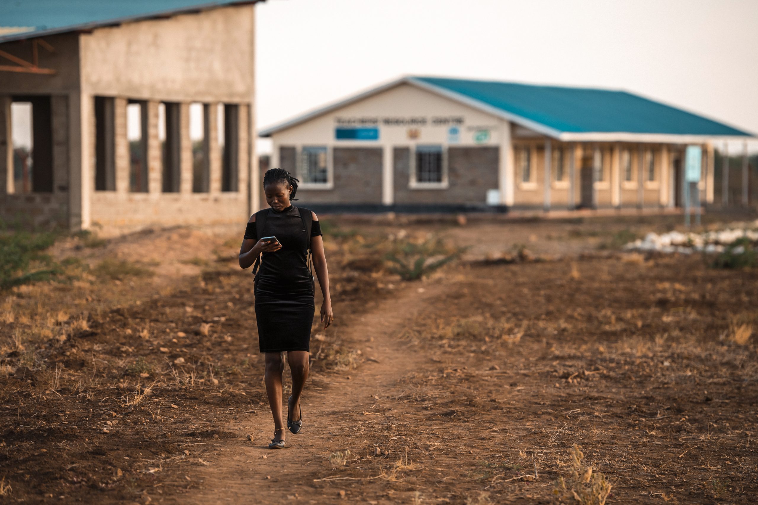 Nainen kävelee ja lukee matkapuhelimensa näyttöä. Taustalla pakolaisleirin koulurakennuksia.