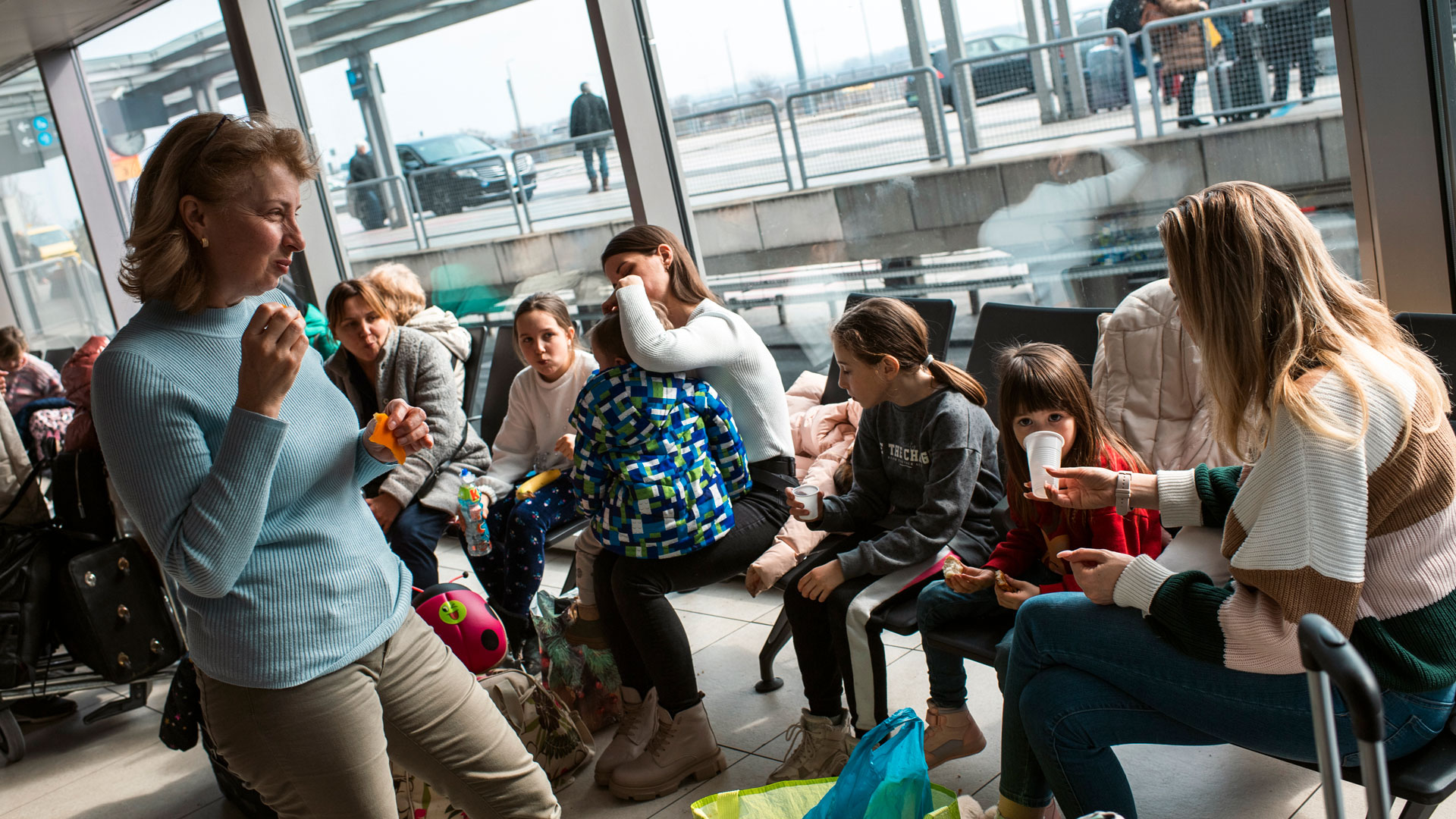Joukko naisia ja lapsia istuu lentokentän isojen ikkunoiden edessä.