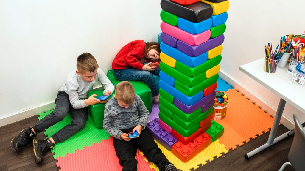 Three children sit on a floor looking at their smartphones. There is a giant Lego tower next to children.