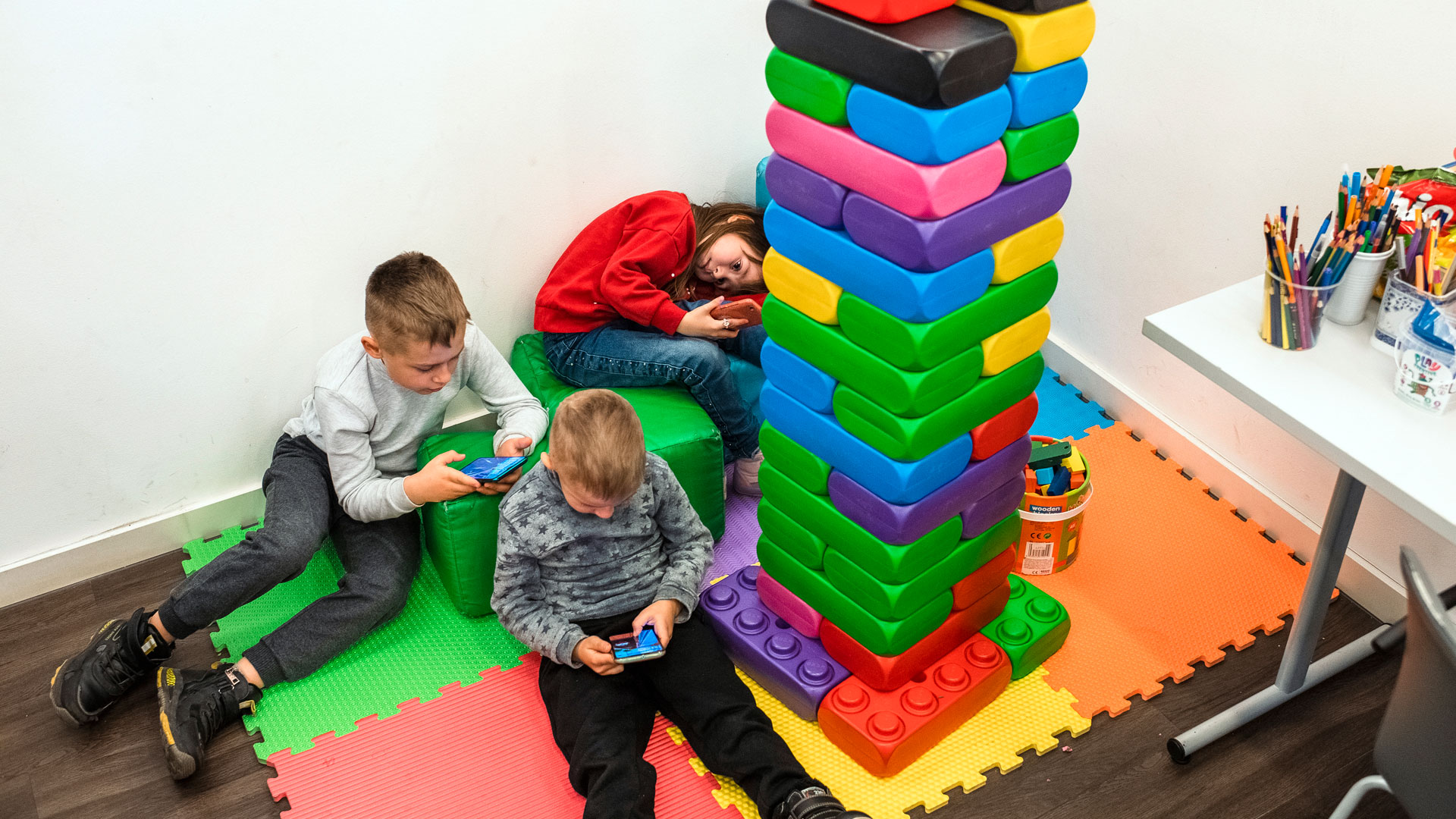 Three children sit on a floor looking at their smartphones. There is a giant Lego tower next to children.