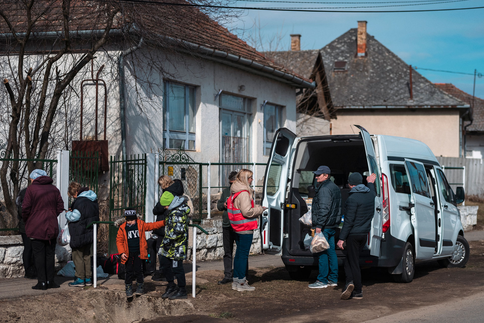 Katunäkymä. Tien reunassa on pysäköitynä auto, jonka vieressä seisoo ihmisiä.