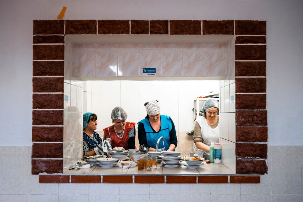 Neljä naista kuvattuna seinässä olevan ikkunan läpi. Naiset työskentelevät keittiössä.