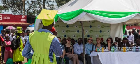 An FCA graduate with his back to the camera stands in green graduation gown and cap while making a speech to an audience at an outdoor graduation ceremony in Kampala, Uganda.