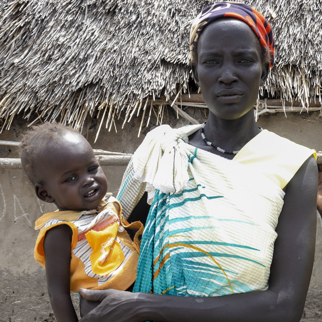 A woman holds a baby in front of a hut in New Fangak, South Sudan