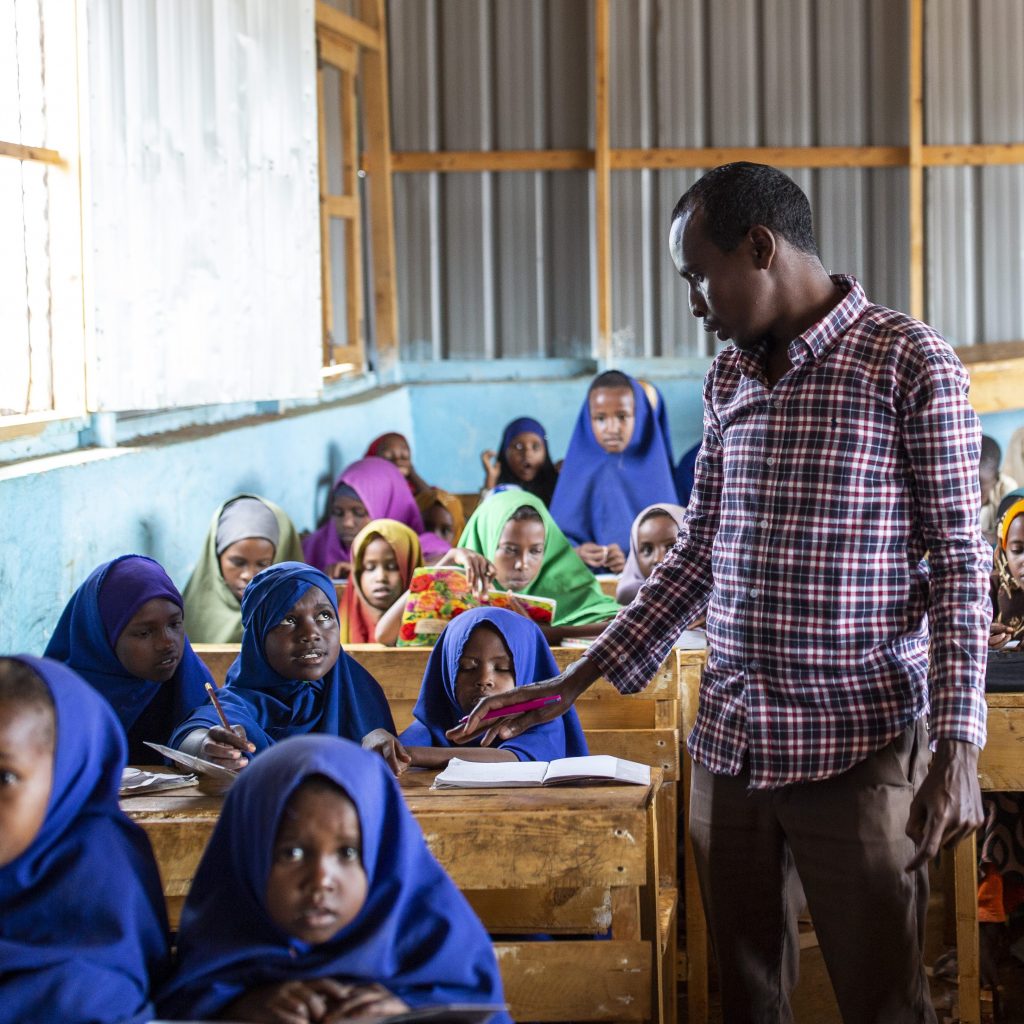 A male teacher standing in a classroom. Lot of students are sitting in behind their desks around the teacher.