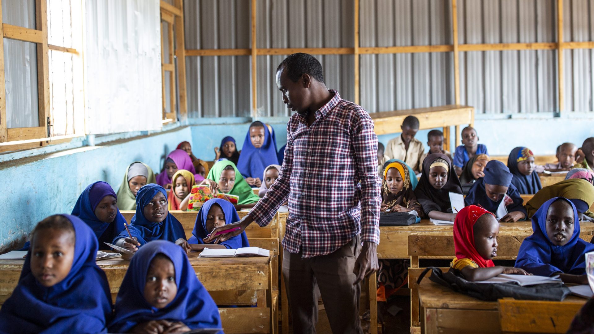 A male teacher standing in a classroom. Lot of students are sitting in behind their desks around the teacher.