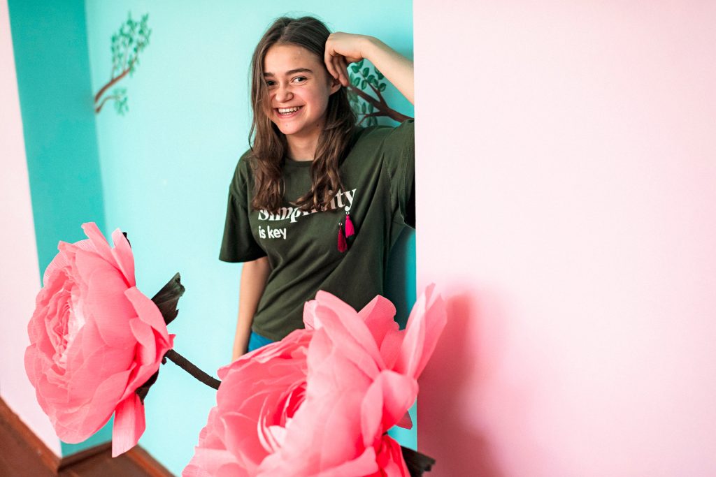 A teenage girl standing next to a wall. Pink roses in front of the girl. Photo: Antti Yrjönen / FCA