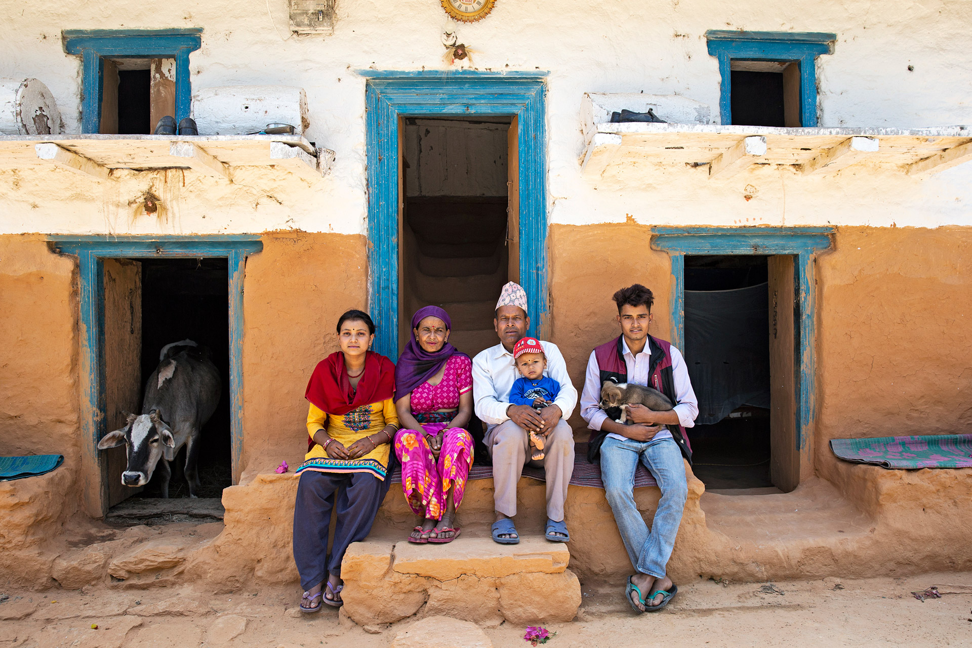 A family is sitting on the porch of their home. A cow is peeking from one of the doorways.