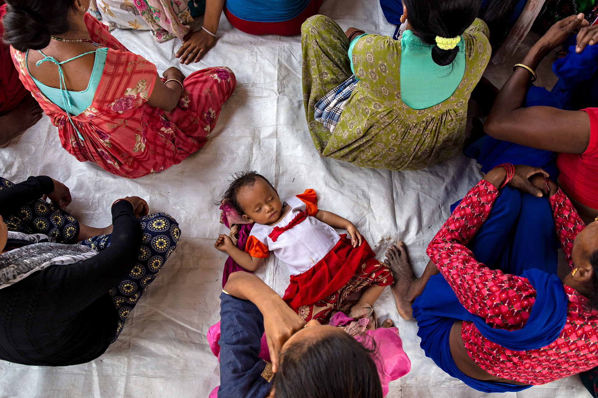 A baby is sleeping on the ground in Nepal. Women sit around the baby.