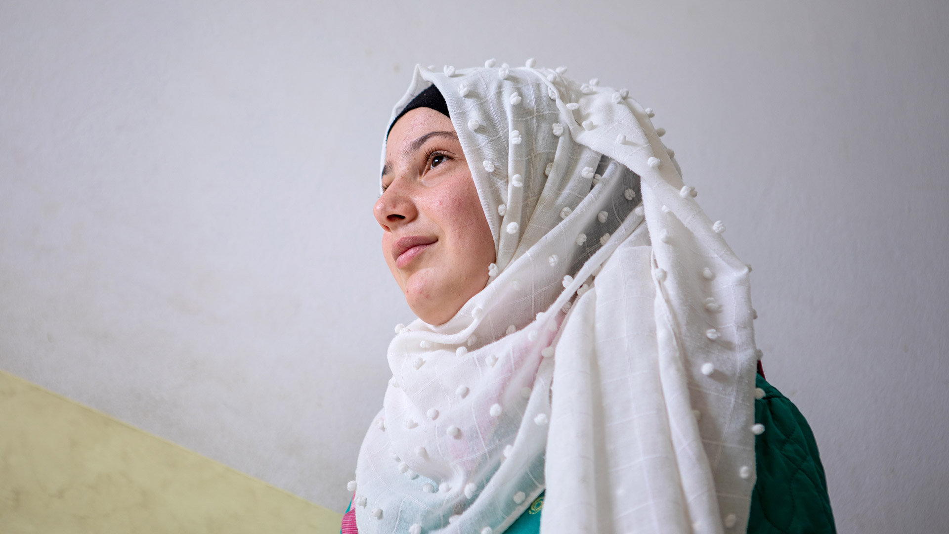 A Syrian girl is photographed from her side in front of a white wall. 