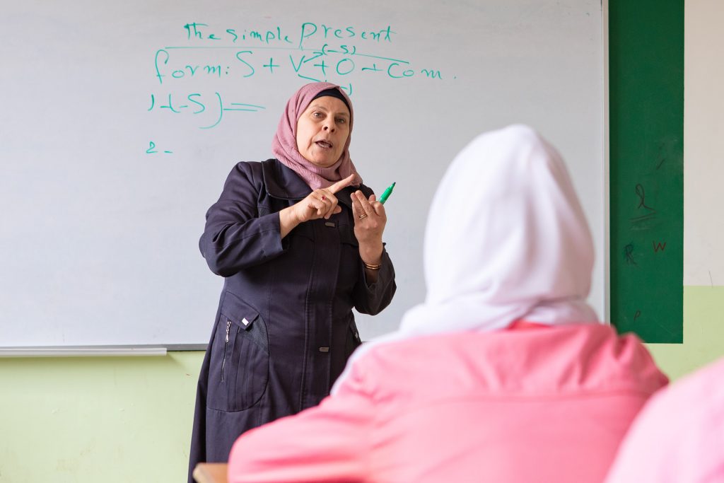 A female teacher is standing in front of her class in Syria. In front of the picture is a girl, photographed from behind.