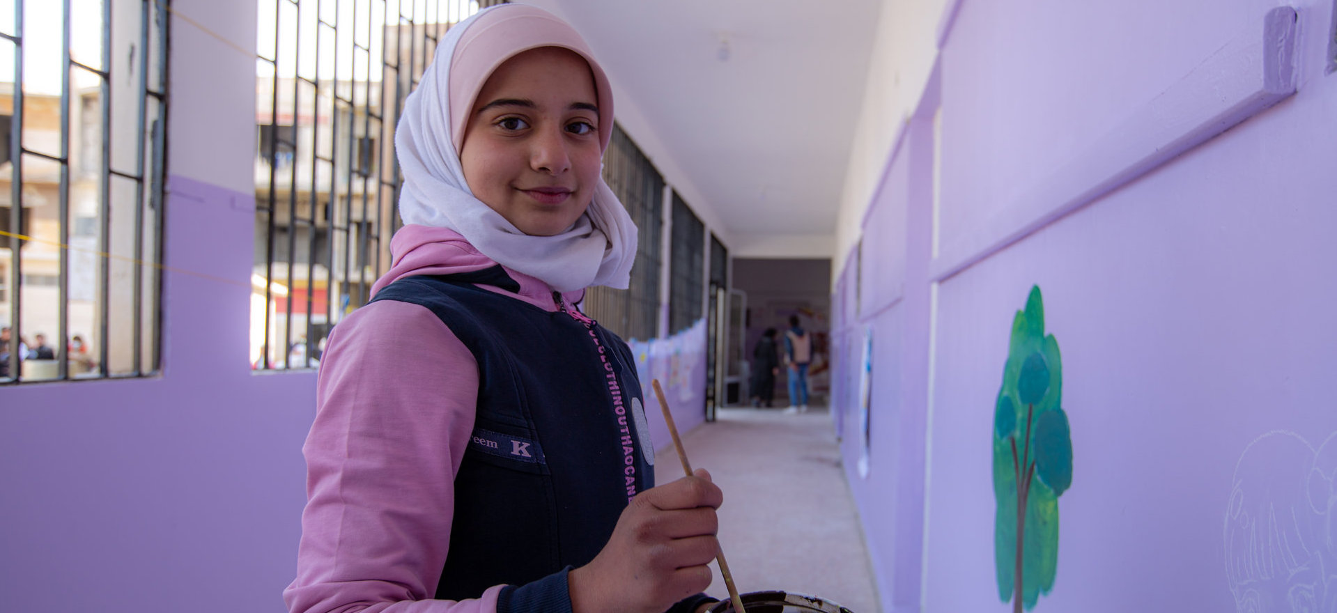 A girl stands in a school corridor in front of a wall with an in-progress mural on it with a paintbrush in her hand.