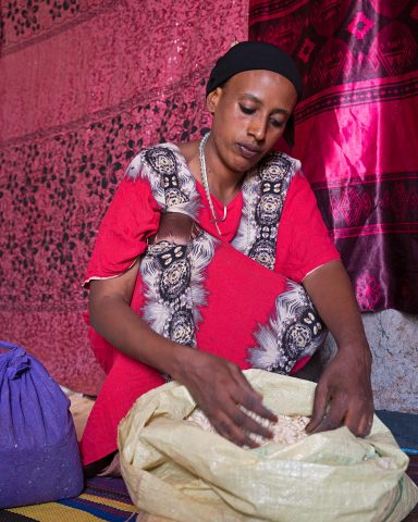 A Kenyan woman sits in front of fabrics. The woman is taking something out from a sack in front of her.