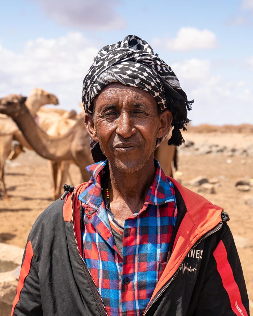 A man wearing a turban looks at the camera. There are camels behind the man. 