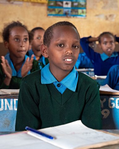 A boy sits behind his desk in a school in Kenya. There is a notebook and a pen on the desk. Other children sit behind the boy.