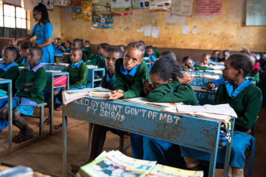 Children sit behind their desks in a class room in Kenya. There are lot of notebooks and papers on the desks.