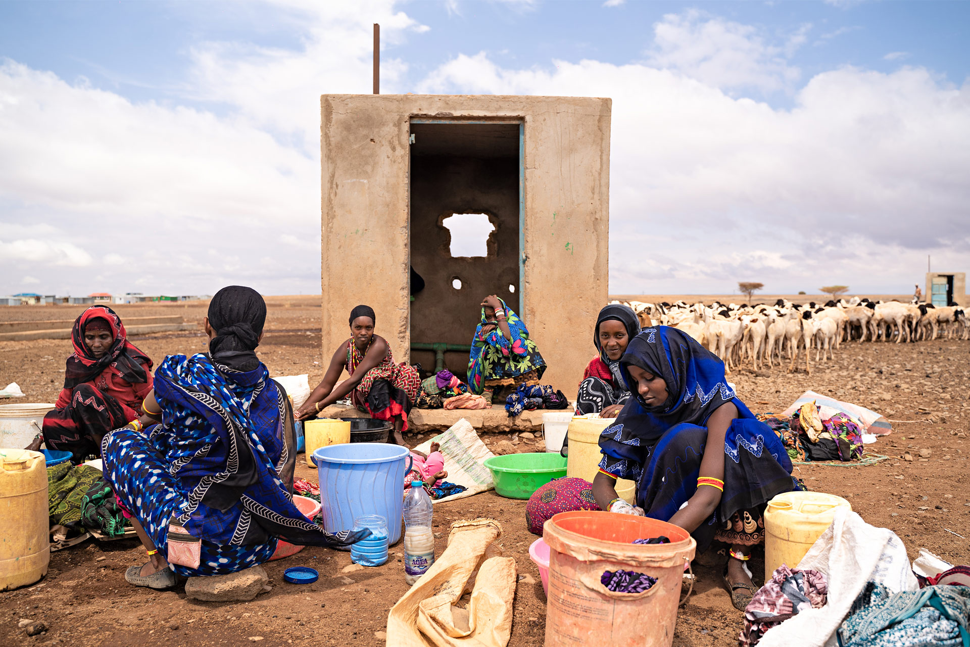 Joukko naisia istuu kuivassa maassa. Naisilla on isoja ämpäreitä. Osa naisista pesee käsin pyykkiä. Taustalla on betonista rakennettu pieni rakennus.