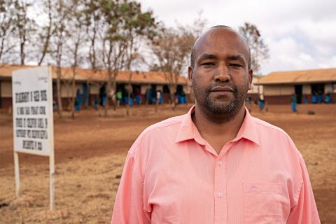 Kenialainen mies katsoo vakavana kameraan. Taustalla näkyy koulurakennus. Vasemmassa reunassa kuvaa on valkoinen kyltti, jossa on mustaa tekstiä. 