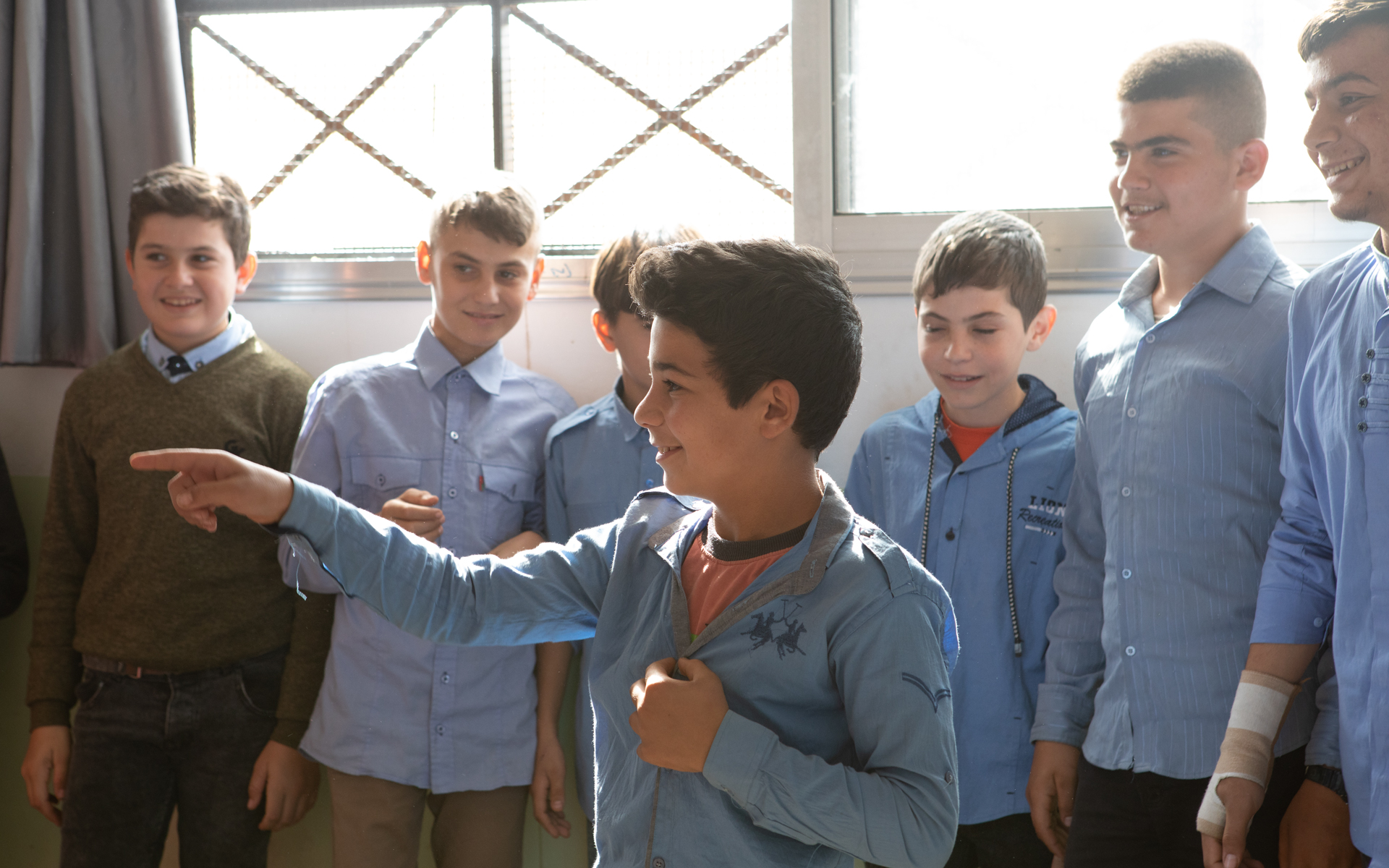 Smiling boy is poking someone with his finger. A group of boys stand behind him in front of a window.