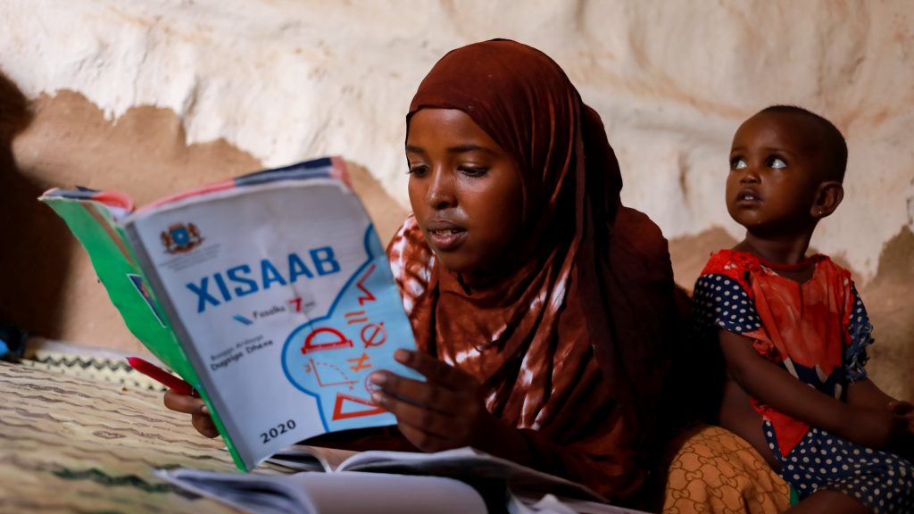 Somalialainen tyttö lukee kirjaa. Taustalla pieni lapsi. 