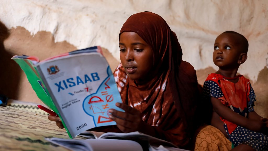 Somalialainen tyttö lukee kirjaa. Taustalla pieni lapsi.