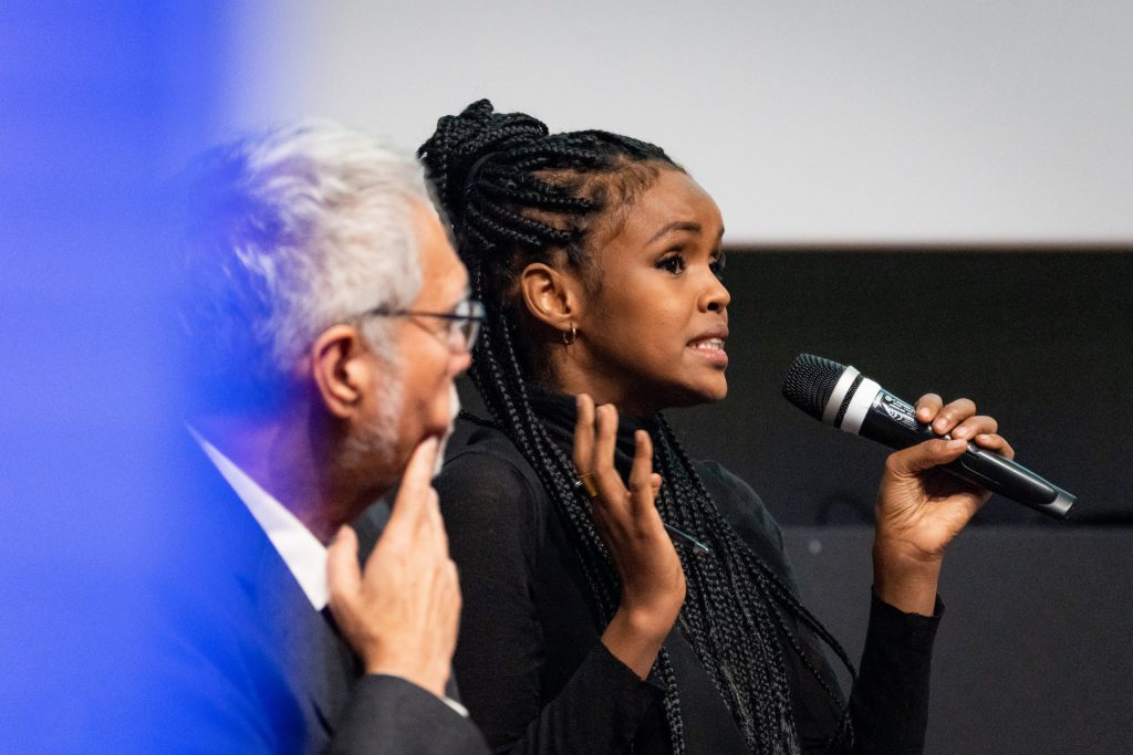 A woman holds a microphone mid-speech. A man listens to the side.