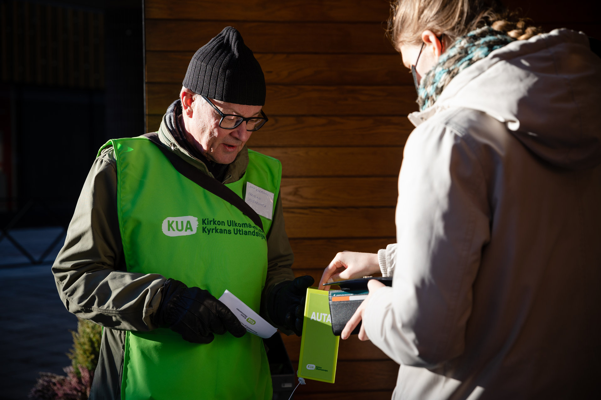 A man in a KUA green vest is collecting money in a box outside in the street. A person is contributing to the collection.