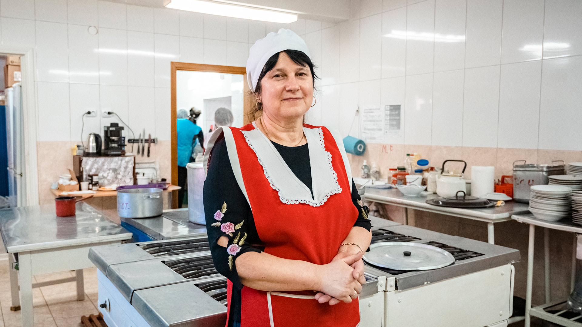 A woman in a cook's outfit clasps her hands and looks at the camera while standing in the middle of a large kitchen