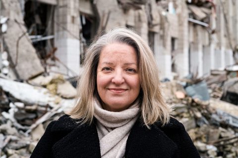 Nainen hymyilee kameralle Ukrainassa. Naisen takana näkyy raunioita.