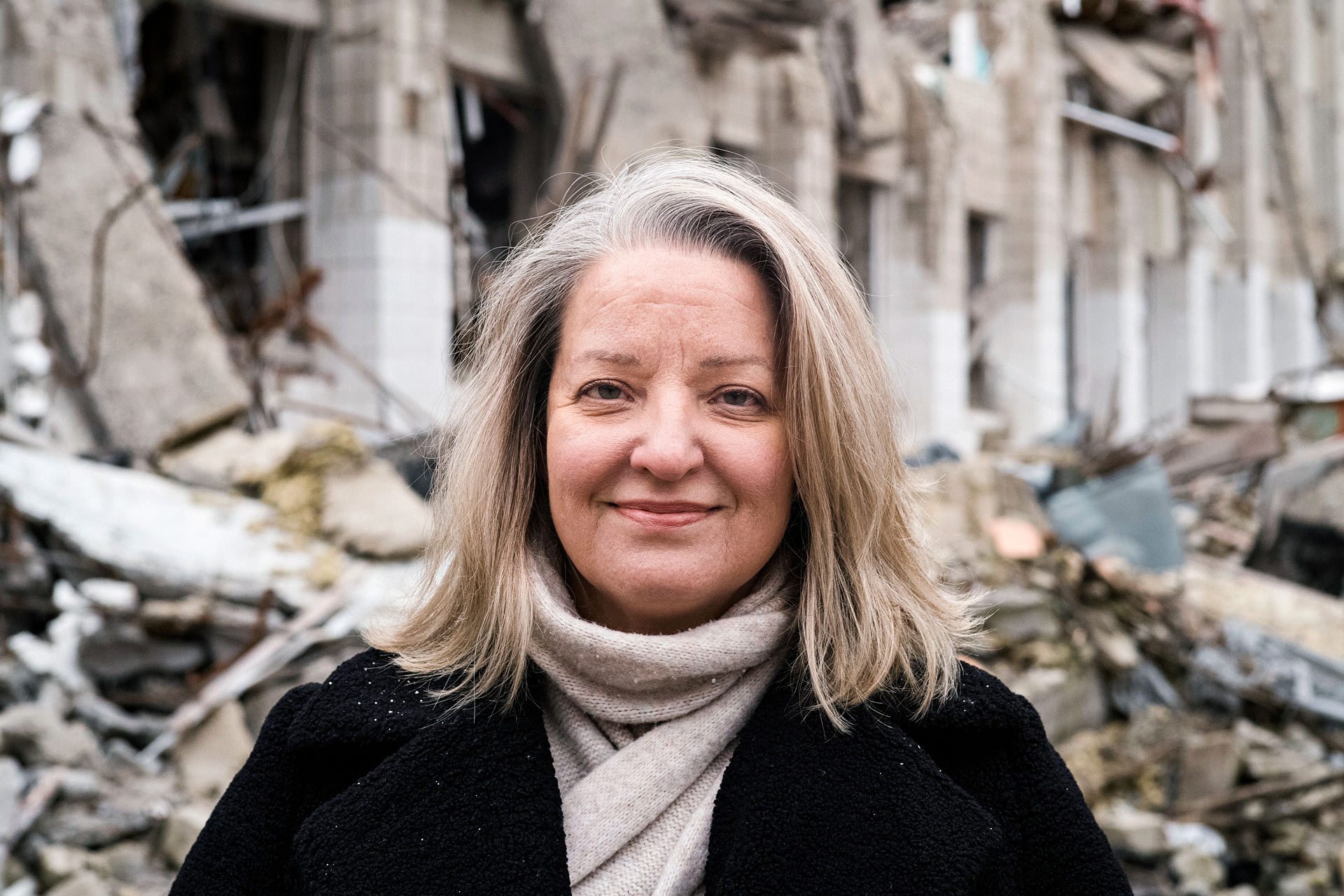 A woman standing in front of a destroyed building smiles at the camera.