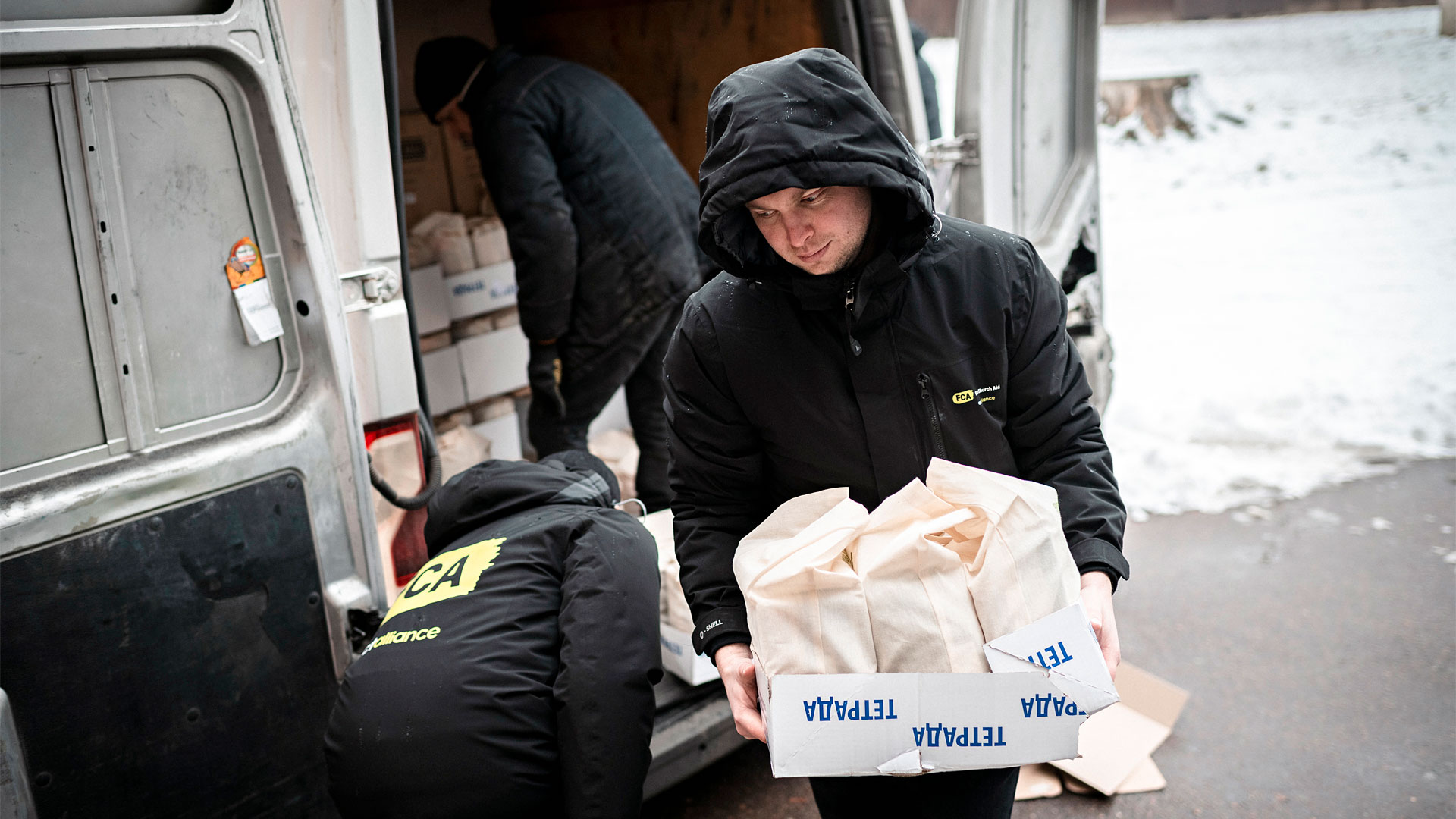 A person in a hooded winter jacket unloads bags of items from a packed van. The jacket bears the logo of FCA.