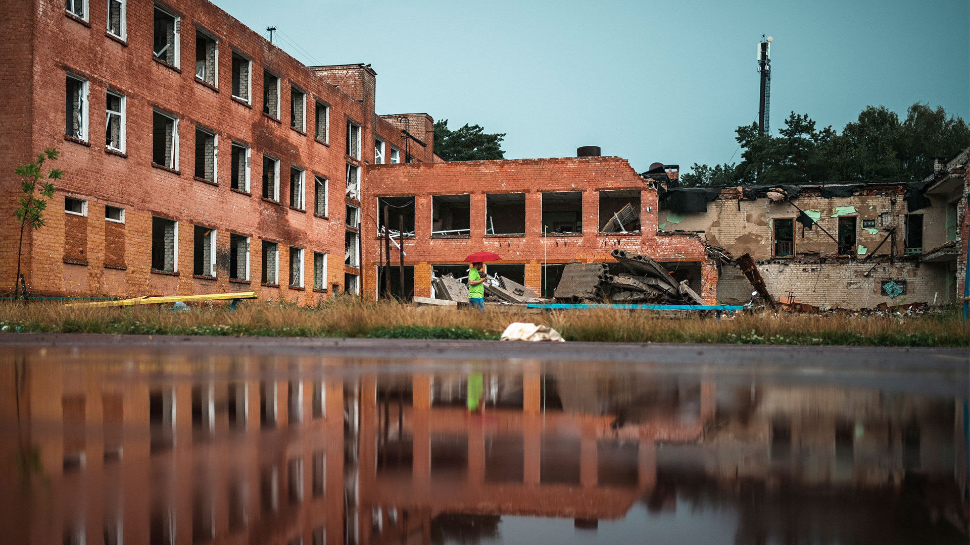 A destroyed school is depicted on the other side of a reflective pool of water. In front of a school a person in a green t shirt holds a red umbrella.