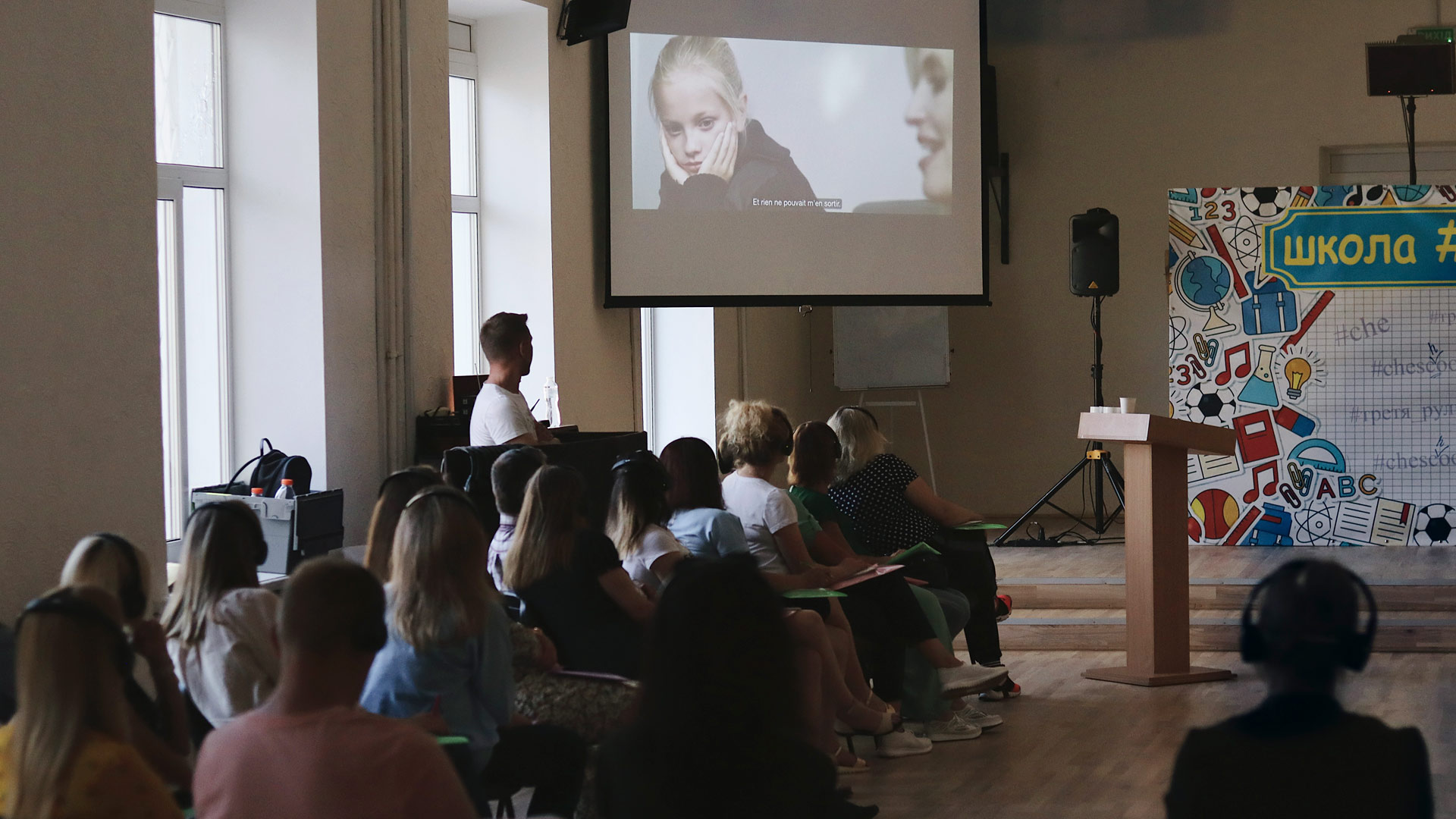 A group of people are seated in an auditorium looking at a screen. The screen has a child leaning on her hands listening to an adult talk.