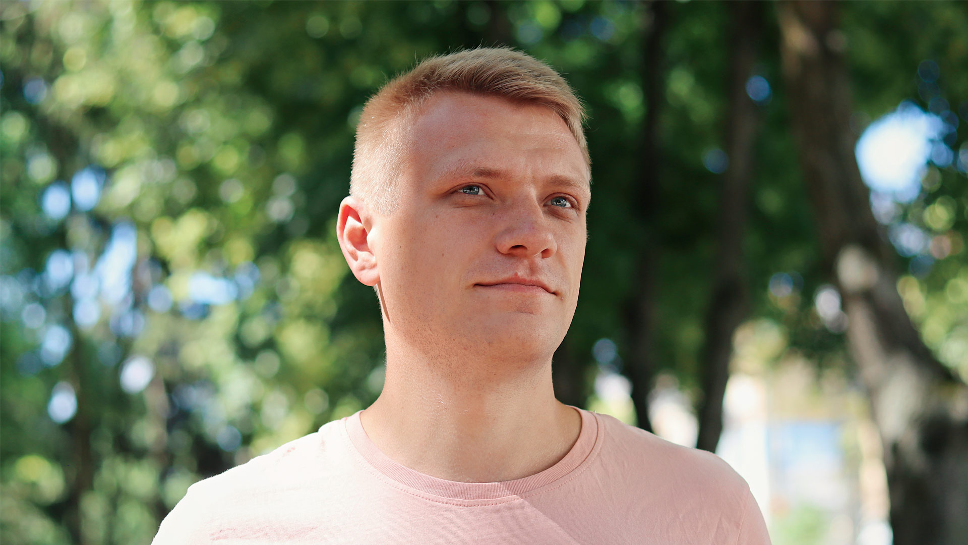 A man looks slightly off-camera in front of a background of sunny trees.