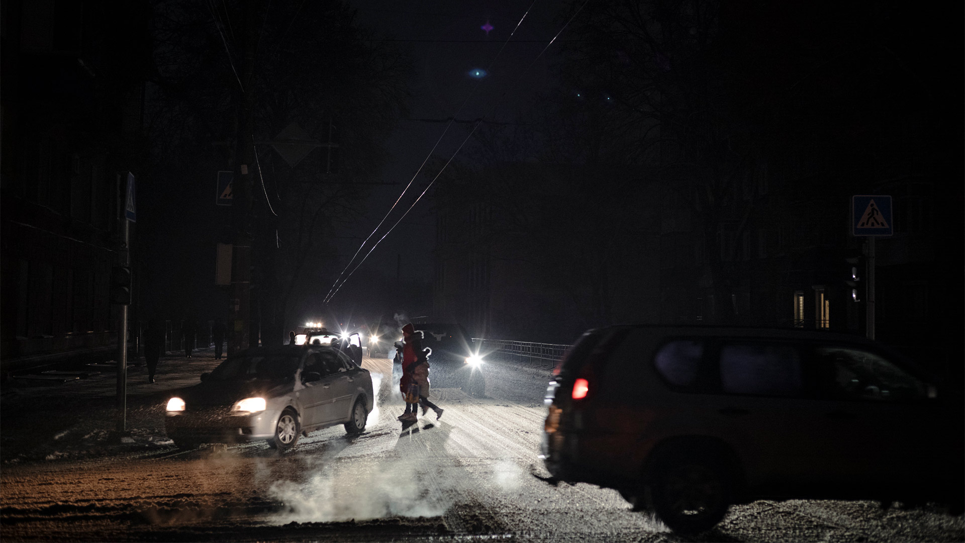 A dark street is only lit up by the light from car headlamps. People cross the street lit up by the headlamps.
