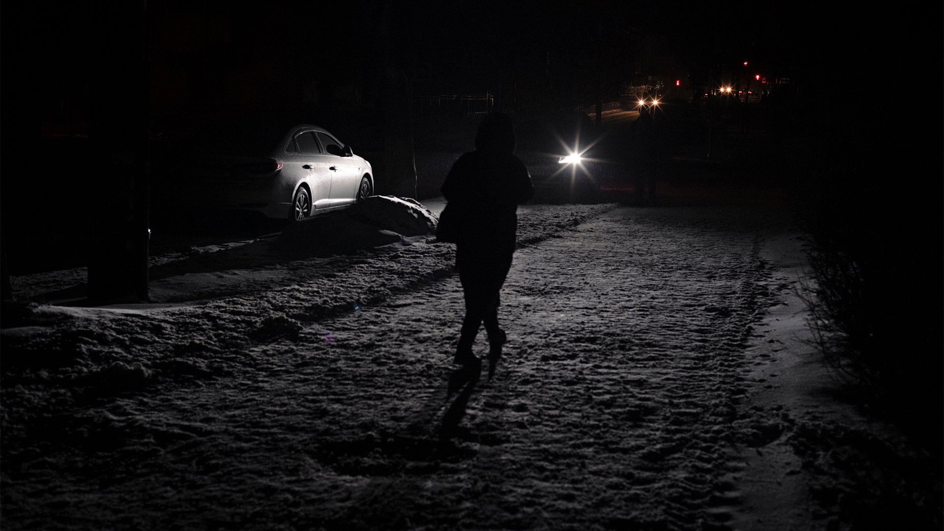 A dark street with a person sillohuetted is only lit up by the light from car headlamps.