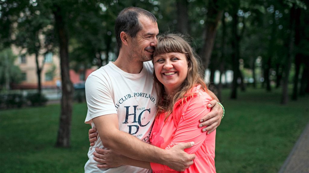 A man and a woman stand in a park with their arms around each other.