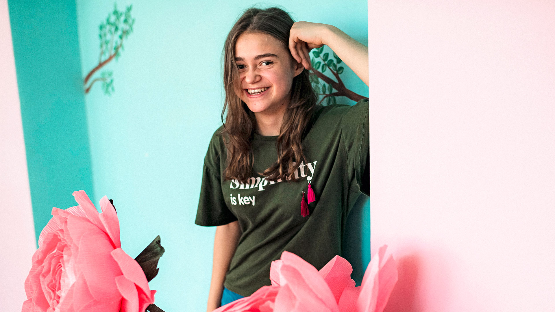 A teenaged girl laughs as she leans against a brightly coloured wall. Pink flowers are in the foreground