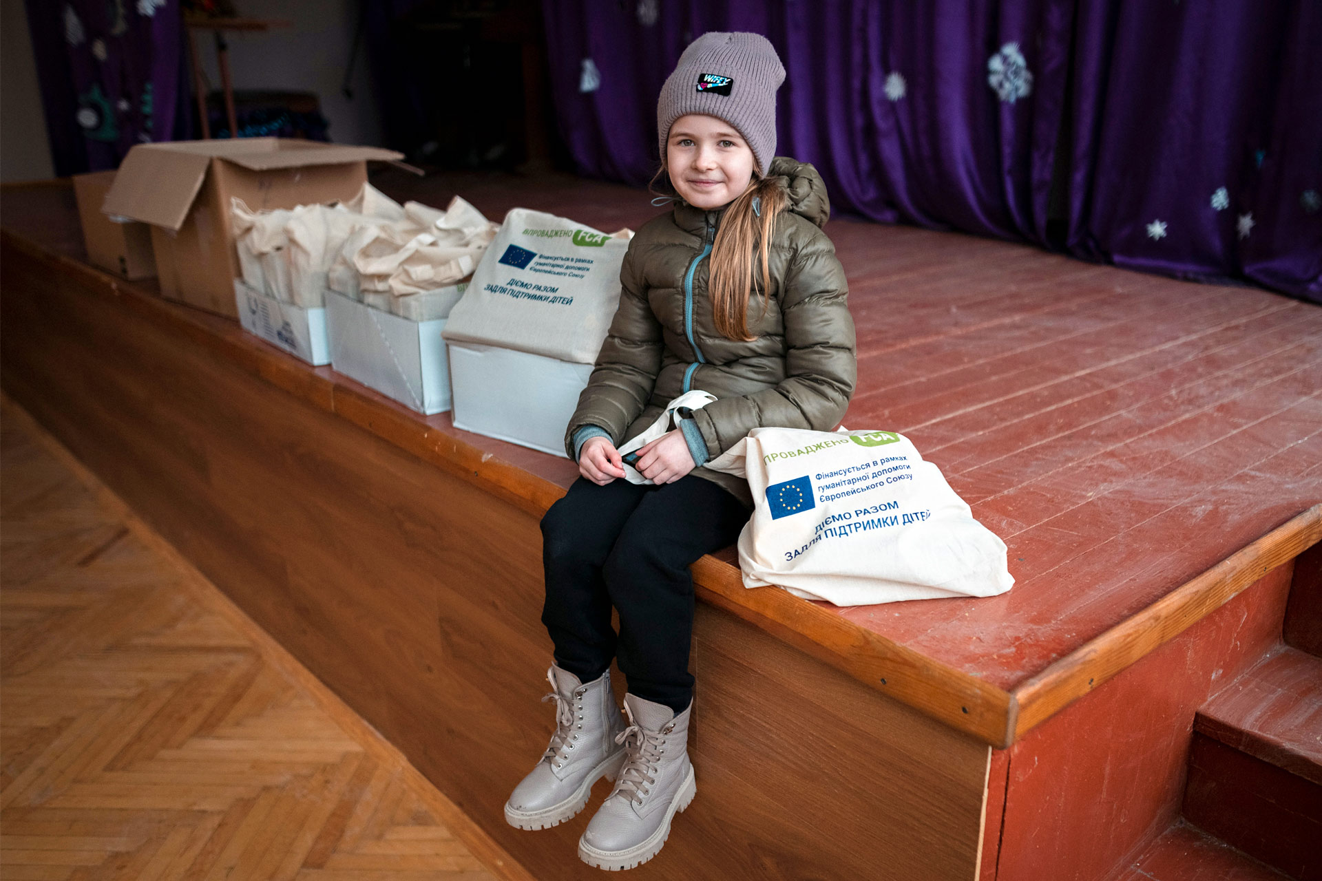 A girl sits on a school stage next to bags full of books that bear the logos of FCA and the EU