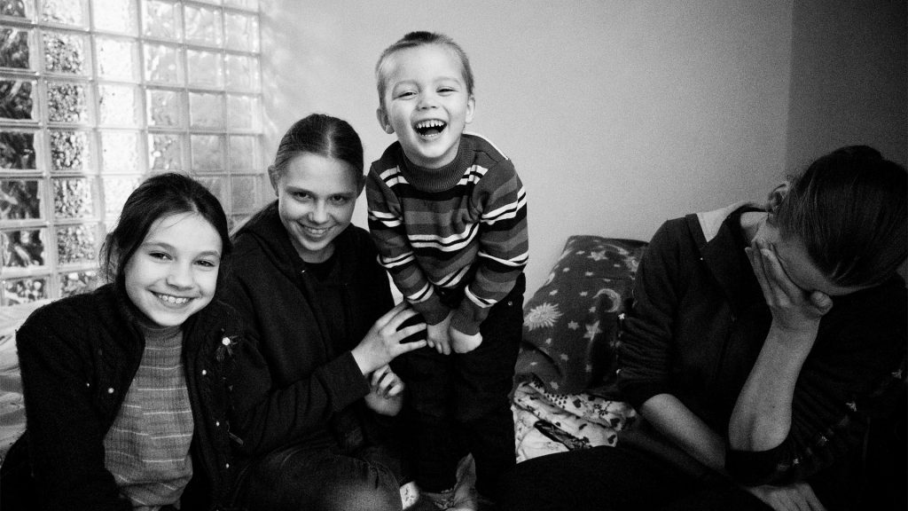 Two girls and a small boy on a bed smile at the camera. An adult woman sits to the side with her hand over her eyes.