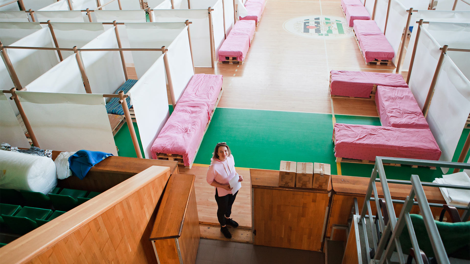 A makeshift shelter in a gym is viewed from above. A woman stands at the entrance holding a piece of paper.