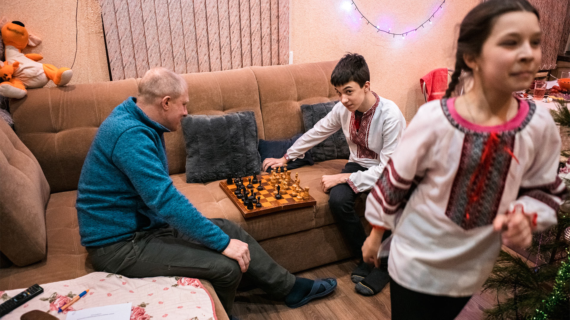 A man and a teenaged boy sit on a sofa with a chessboard between them. A girl in the foreground walks out of the shot.