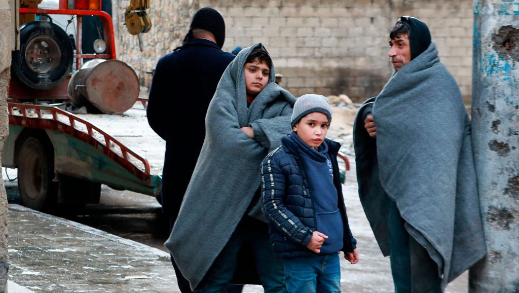 Two Syrian boys and a man wrapped in blankets stand outside. 