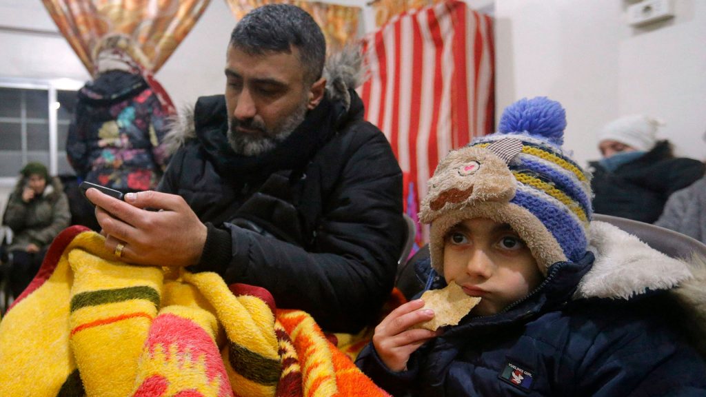 A Syrian child sits wrapped in a blanket eating a cookie. There is an older man sitting behind the child.