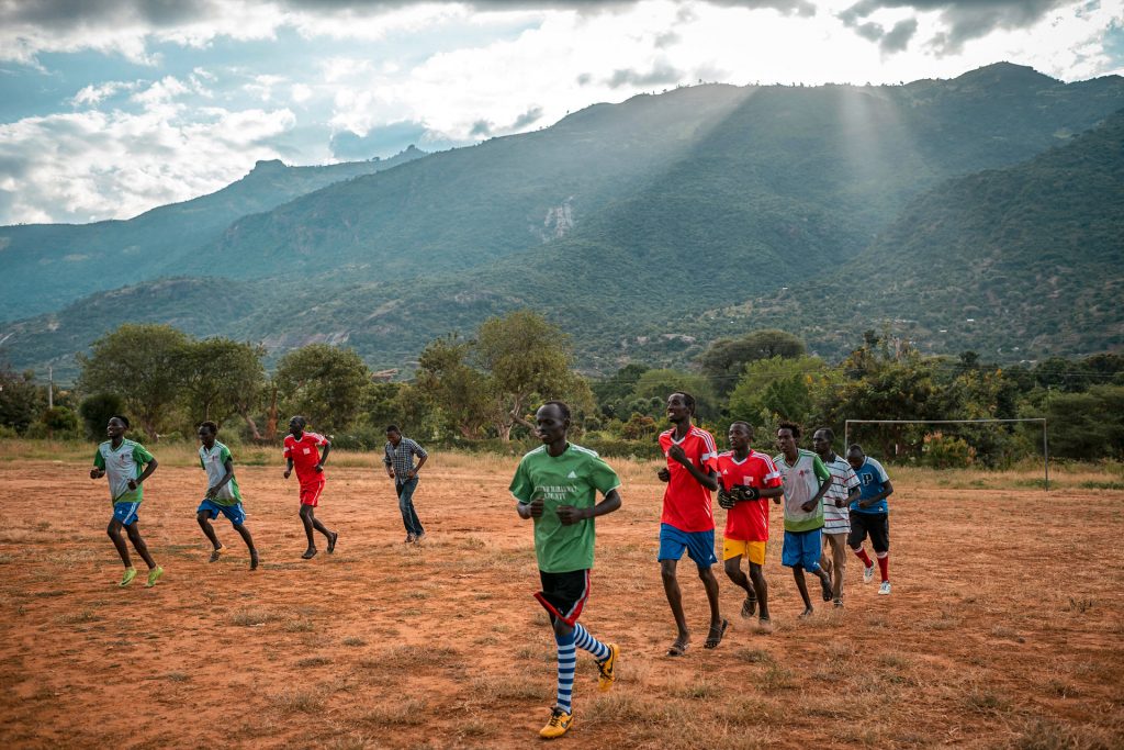 Jalkapallon peliasuihin pukeutuneet kenialaiset miehet hölkkäävät hiekkakentällä. Taustalla näkyy korkea kukkula.
