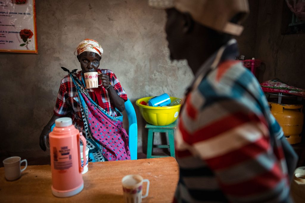 Kenialainen nainen istuu seinän edessä juomassa teetä. Pöydällä on termoskannu. Kuvan etualalla on selin kameraan oleva mies. 