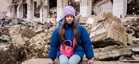 A teenaged girl in a blue winter jacket, a purple hat and holding a pink bag sits in front of the rubble of a destroyed school.