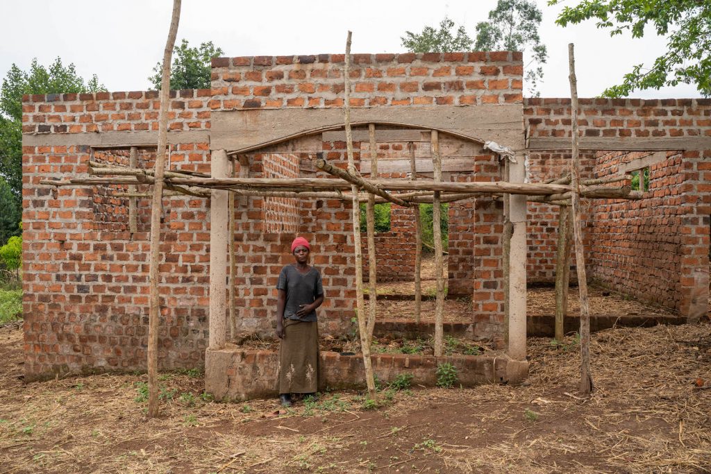 A Ugandan woman standing in front of a brick house under construction, the walls are up, but the roof is missing.